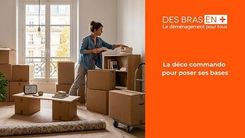 Une femme dans un appartement rempli de cartons de déménagement place des livres sur une étagère, avec une vue sur des bâtiments parisiens par une fenêtre.